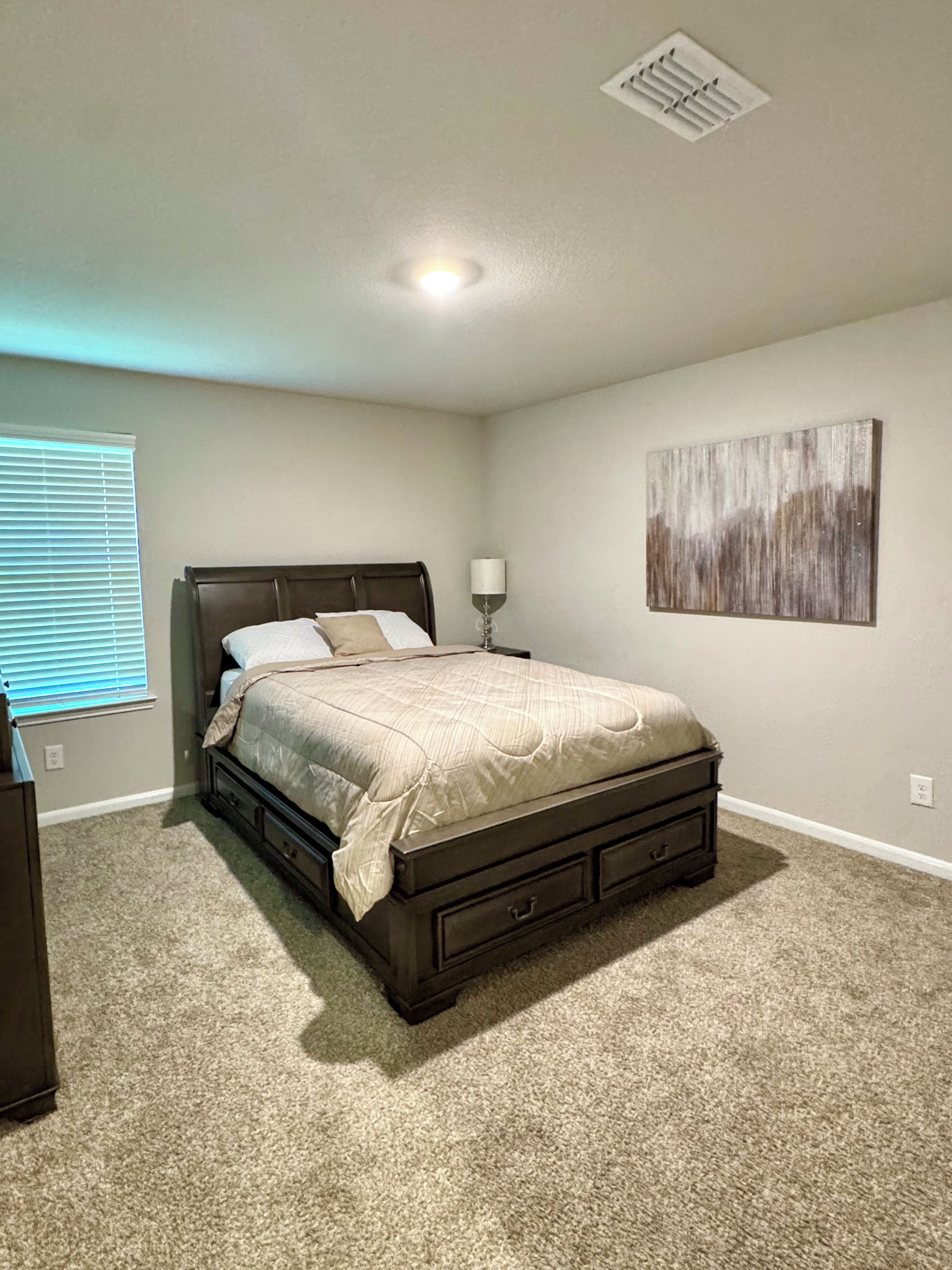 Une chambre à coucher avec un lit, une table de chevet, une lampe, un tableau au mur et une bouche d’aération au plafond.