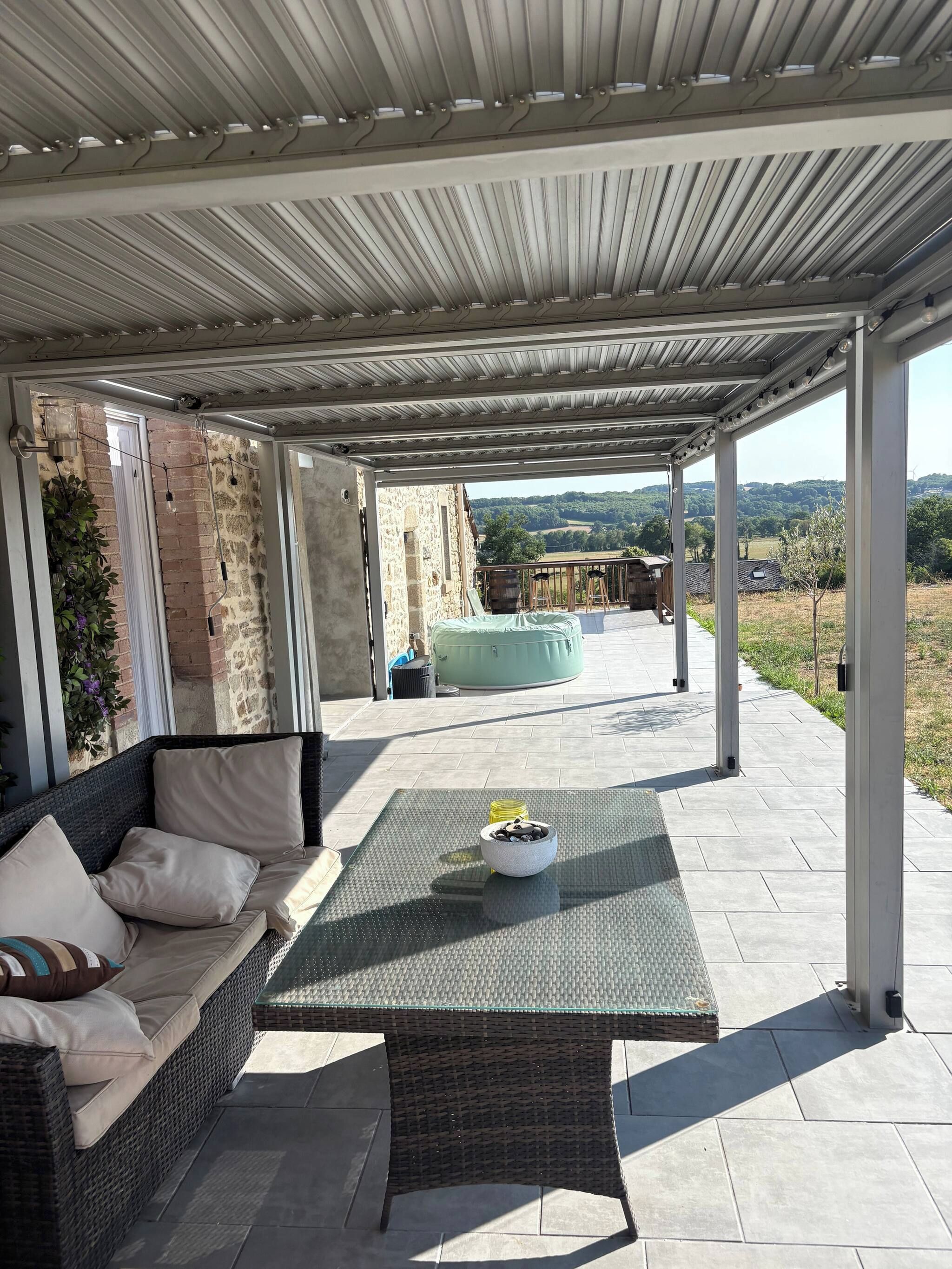 A covered outdoor seating area with a wicker sofa, a table with a bowl of fruit, and a view of a stone building and landscape.