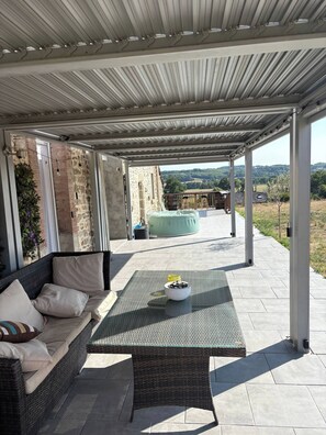 A covered outdoor seating area with a wicker sofa, a table with a bowl of fruit, and a view of a stone building and landscape. - 4 guests with beautiful views (Saint-Agnant-de-Versillat)