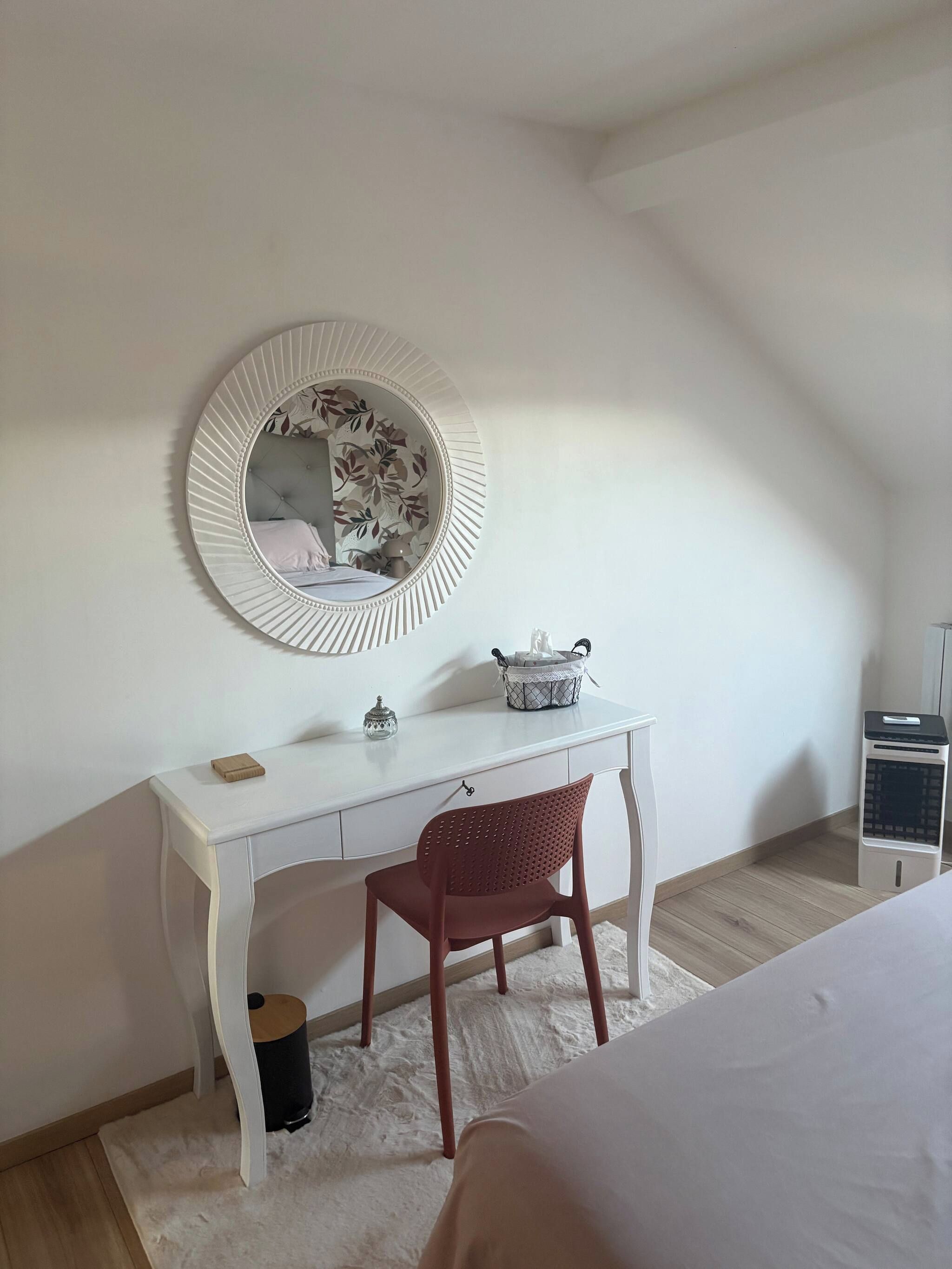 Une chambre avec un bureau blanc, une chaise rouge, un miroir rond et un lit.