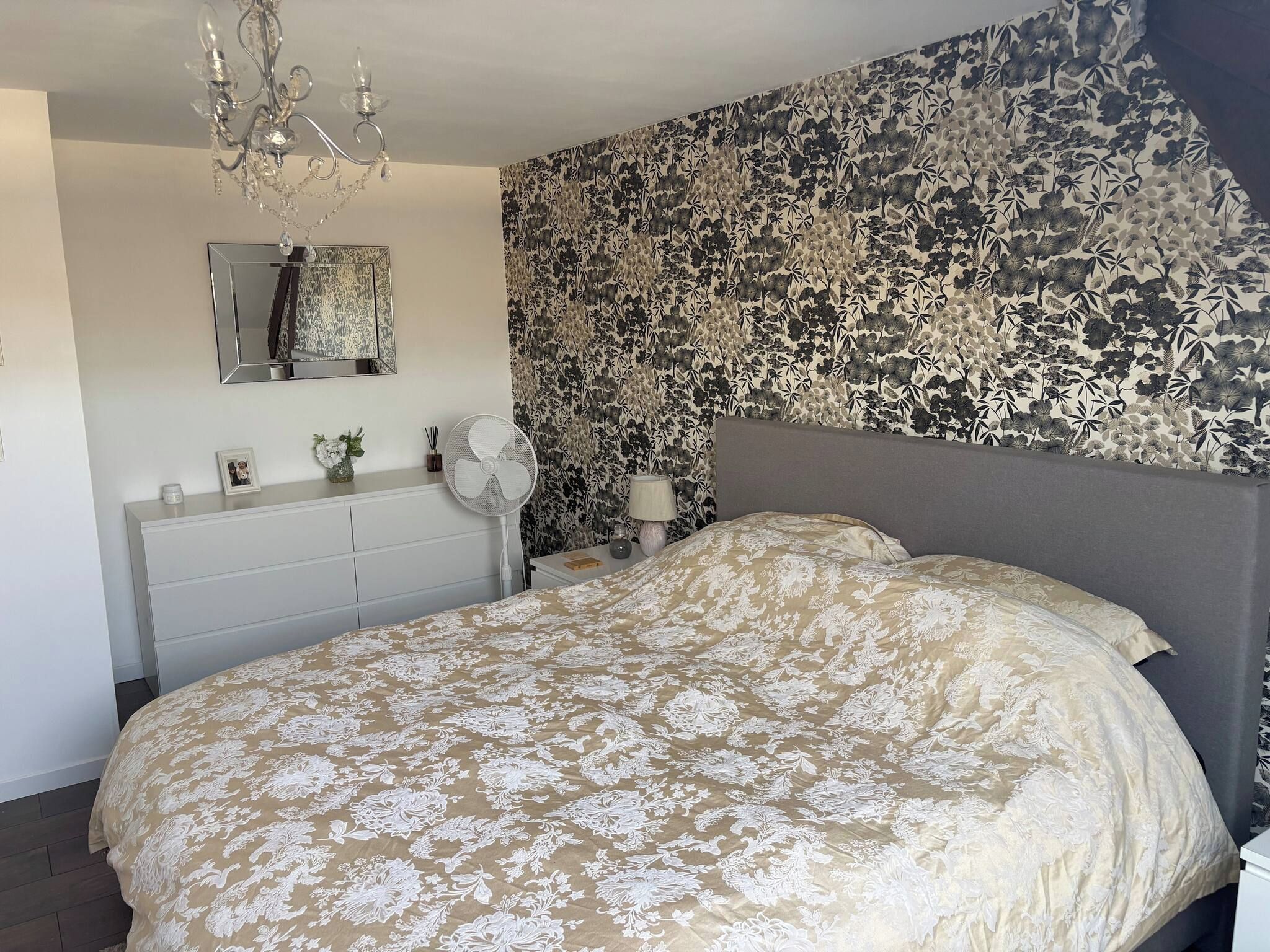 Een slaapkamer met een groot bed, een dressoir, een spiegel, een ventilator en bloemenbehang.