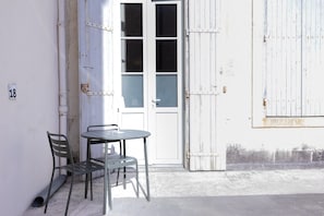 Ein kleiner Außenbereich mit Tisch und zwei Stühlen, eine weiße Tür mit Fenster sowie eine weiße Wand mit einer Nummer.