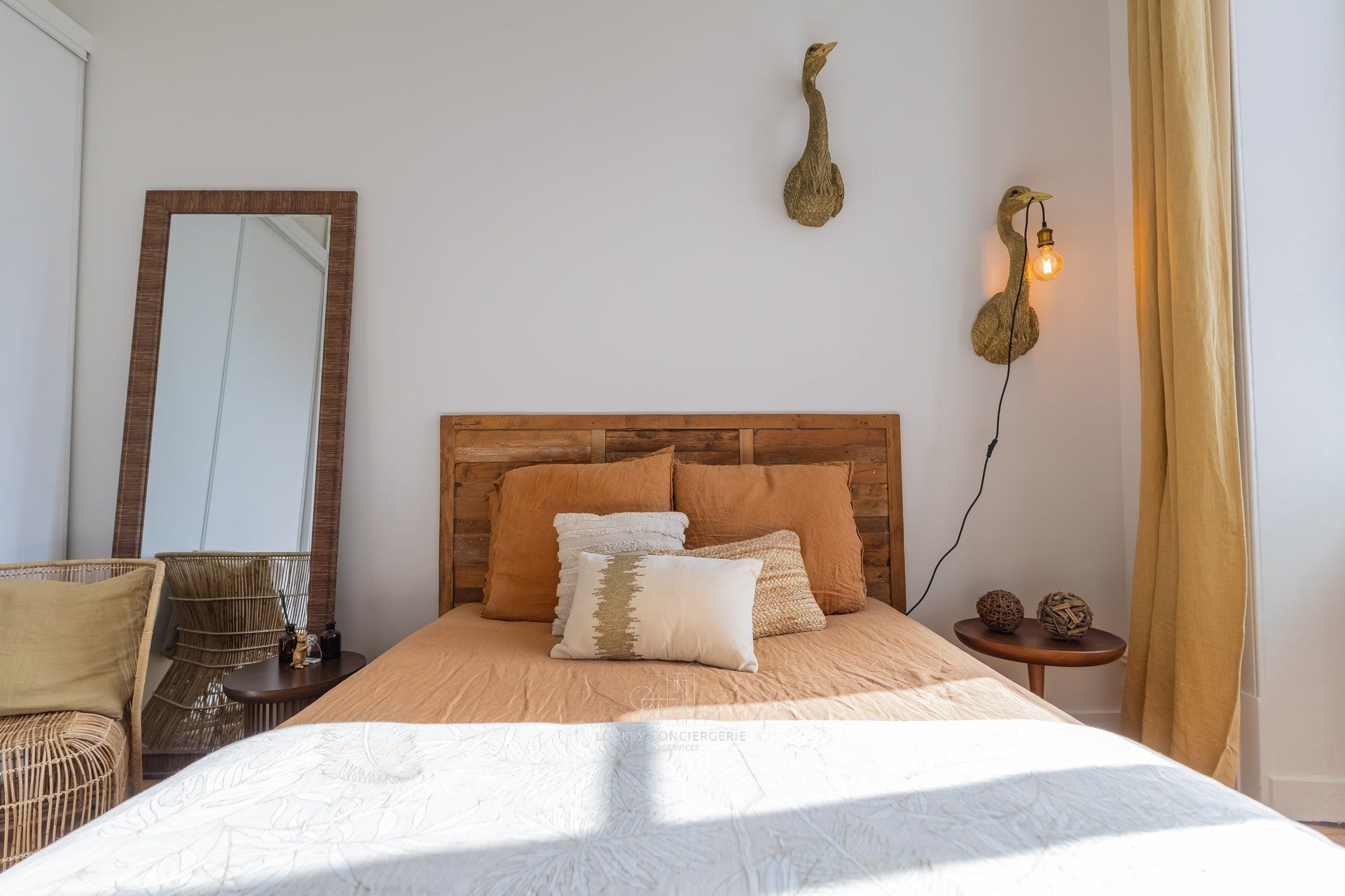 A bedroom with a wooden headboard, a large mirror, a bed with multiple pillows, a wall-mounted light fixture, and a small table with decorative items.