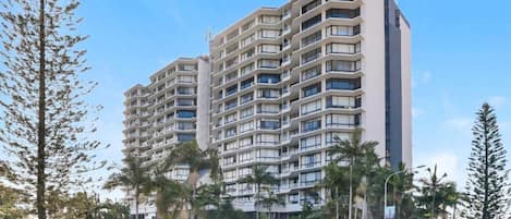 A multi-story residential building with balconies, surrounded by palm trees and a clear sky.