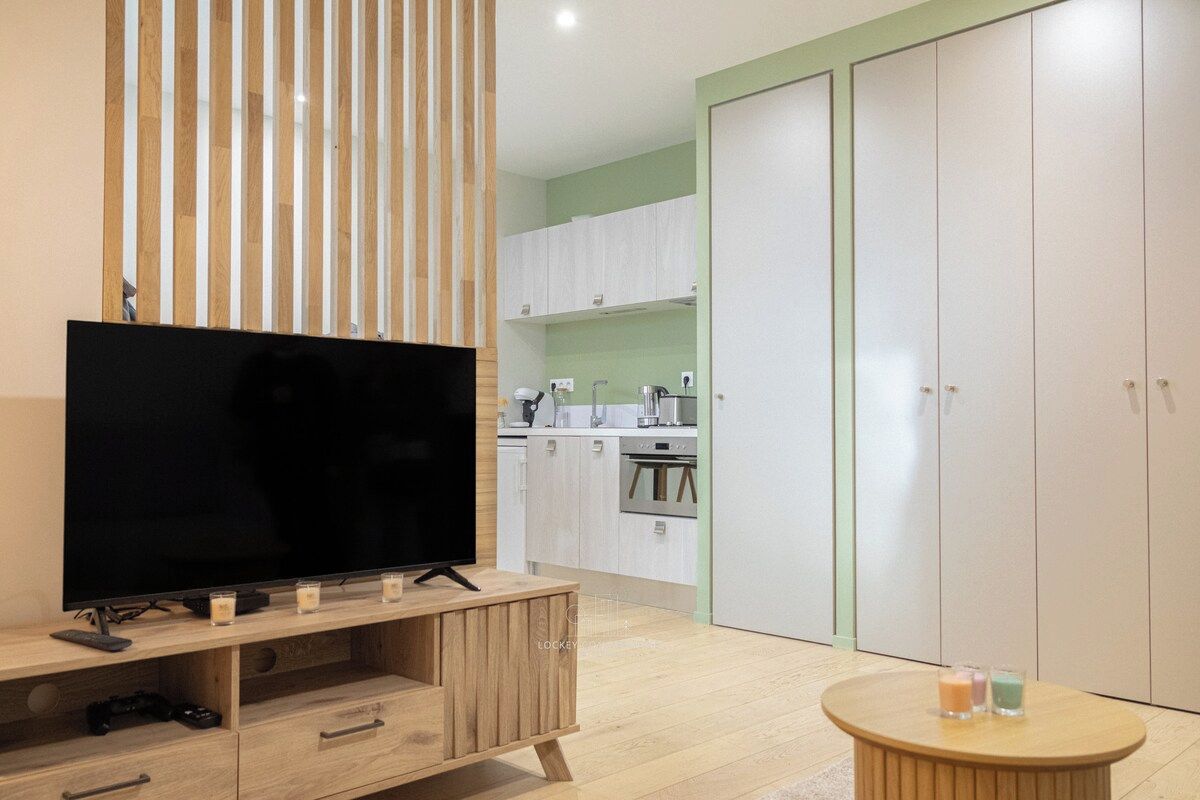 Un salon moderne avec un téléviseur à écran plat posé sur un meuble en bois, une petite table ronde avec des bougies, et une cuisine en arrière-plan avec des meubles blancs et un mur vert.