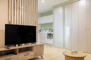 Una sala de estar moderna con un televisor de pantalla plana sobre un mueble de madera, una pequeña mesa redonda con velas, y una cocina al fondo con armarios blancos y una pared verde.