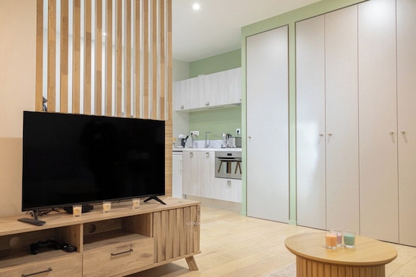 A modern living room with a flat-screen TV on a wooden stand, a small round table with candles, and a kitchen in the background with white cabinets and a green wall. - Joli studio spacieux en hypercentre (La Rochelle)