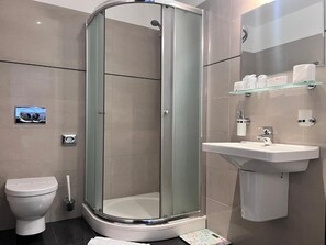 A modern bathroom with a glass-enclosed shower, a toilet, a sink, and a shelf with toiletries. - Hotel TENIS (Zvolen)