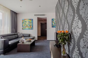 A modern living room with a sofa, coffee table, and a TV on a stand. There are two abstract paintings on the wall and a vase with flowers on a side table. - Hotel TENIS (Zvolen)