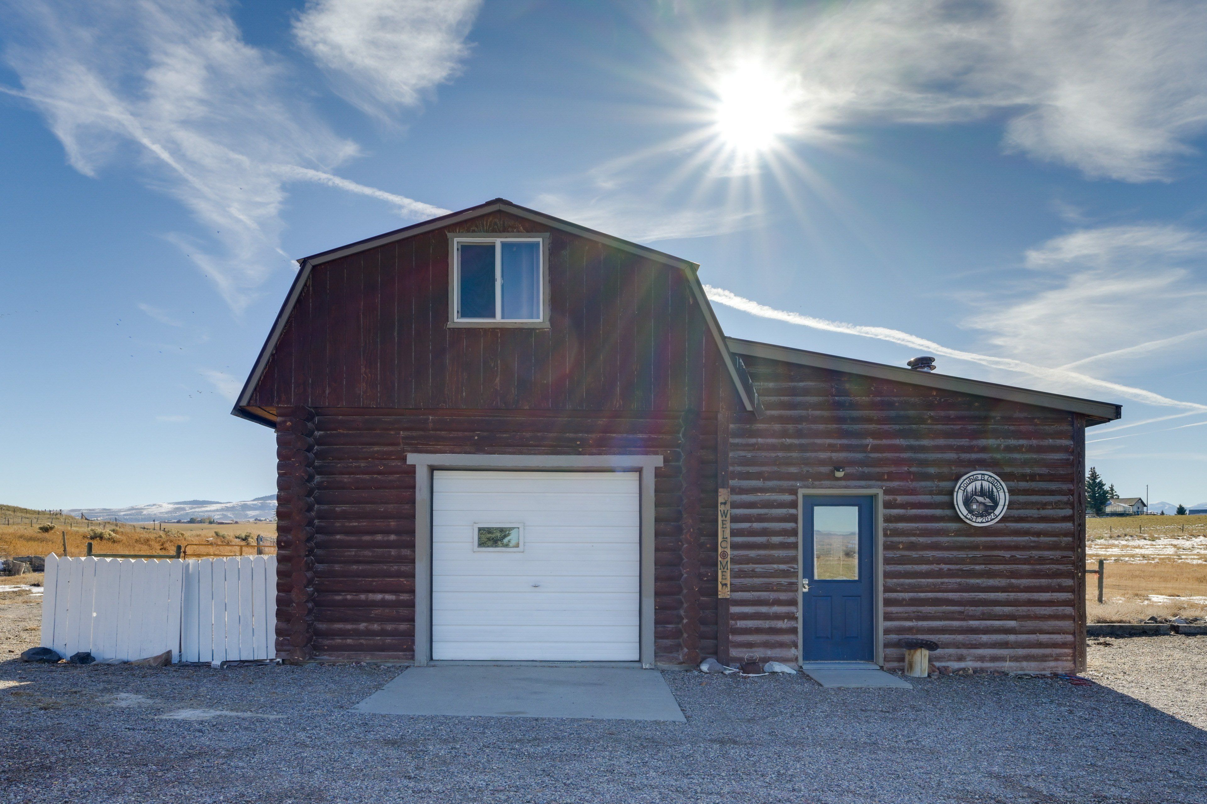 A single-story log cabin with a white garage door, a small window, and a door with a welcome sign.
