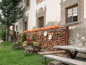 Ein rustikaler Sitzbereich im Freien mit einem Holztisch und Bänken, einem Holzstapel und dekorativen Kürbissen vor einem Gebäude mit verzierten Fenstern.