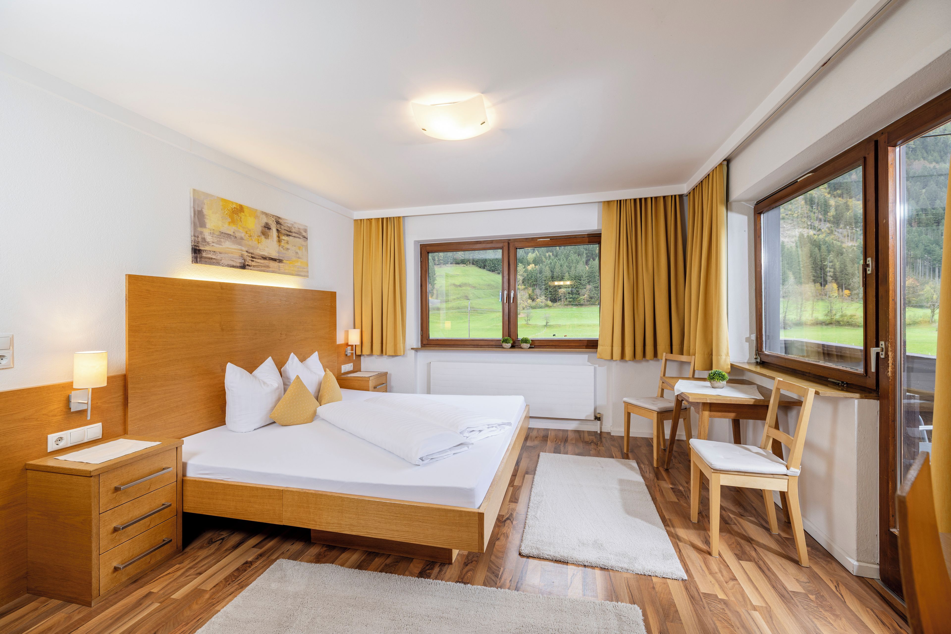 A hotel room with a large bed, wooden headboard, bedside lamps, a desk with a chair, and a window with curtains.