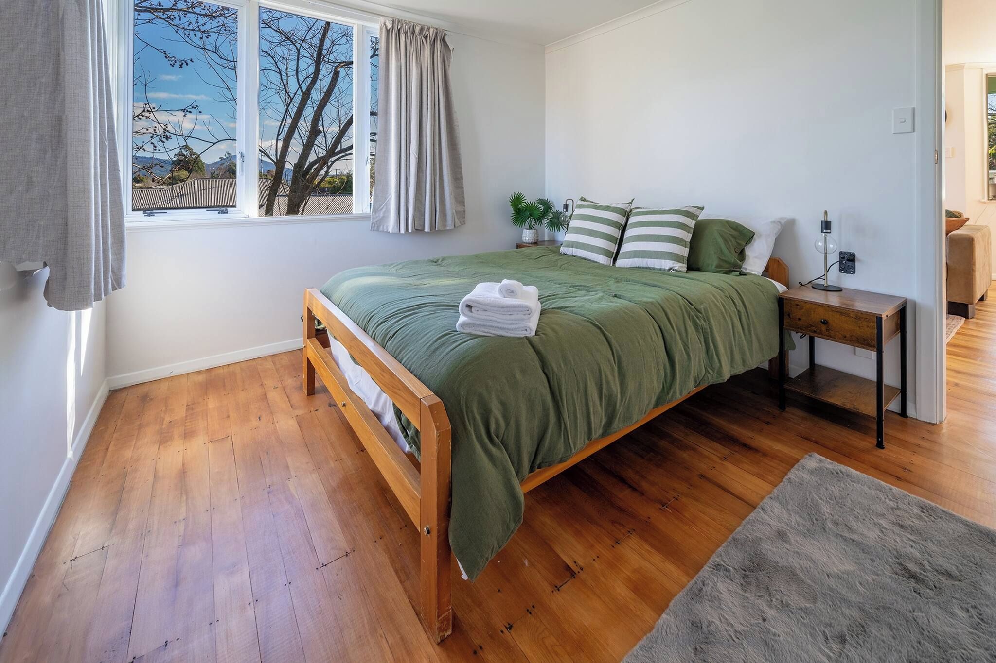 A bedroom with a wooden bed, a bedside table, a lamp, and a view of the outdoors through a window.
