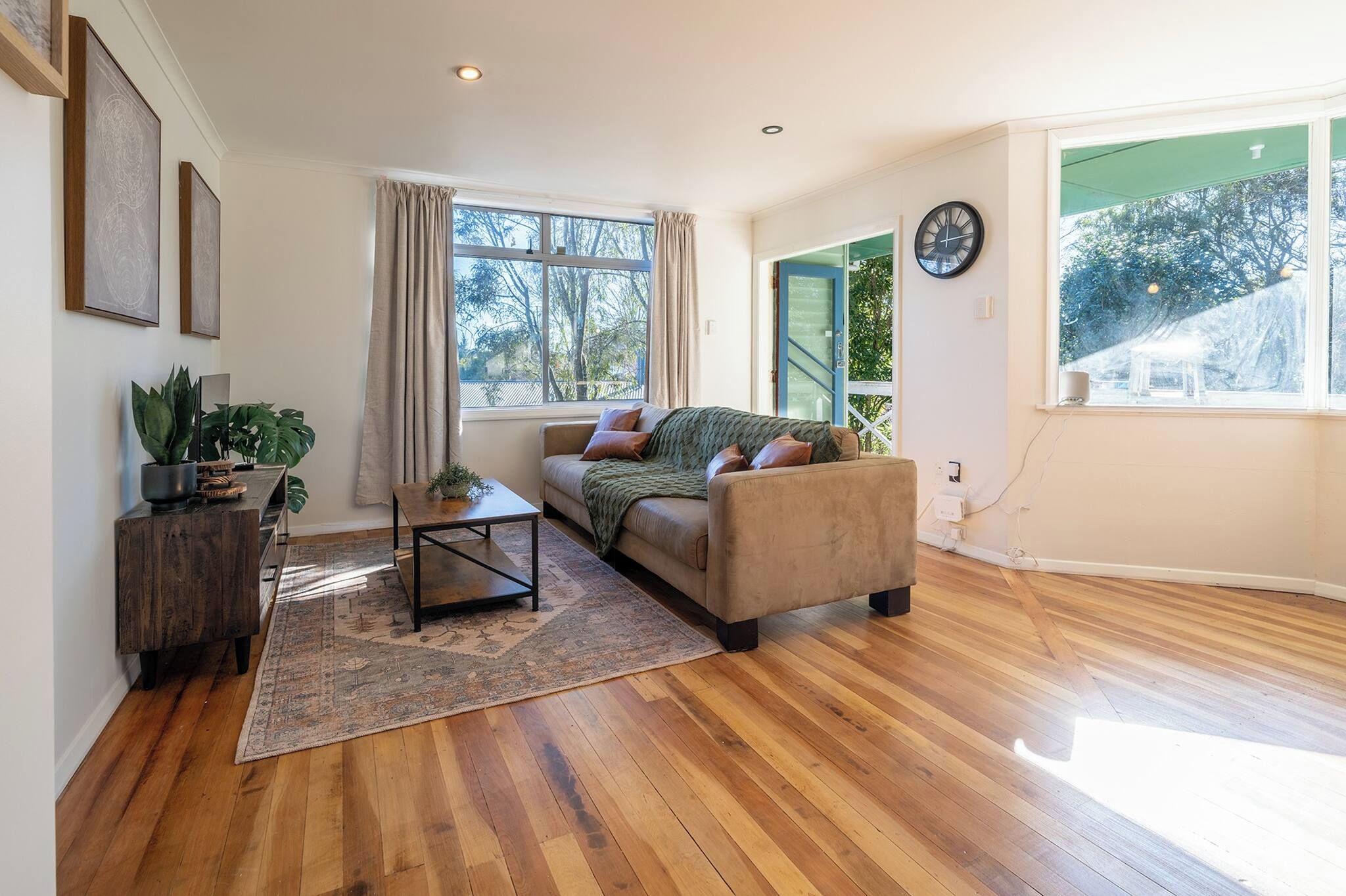 Un salon avec un canapé, une table basse et un tapis. Il y a des fenêtres avec des rideaux, une horloge au mur et une plante sur une table en bois.