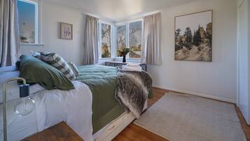 A bedroom with a bed, a bedside table, a lamp, a window with curtains, a painting on the wall, and a rug on the floor.