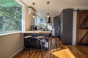 A modern kitchen with a bar area, wooden flooring, and large windows. - The Jumping Trout Rustic Hideaway (Tūrangi)