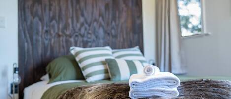 Cama com cabeceira de madeira, travesseiros listrados em verde e branco, e toalha branca enrolada em uma superfície de madeira.