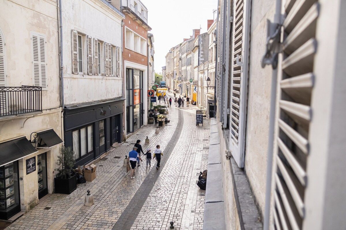 Una estrecha calle adoquinada flanqueada por edificios, algunos con contraventanas, y algunos peatones.
