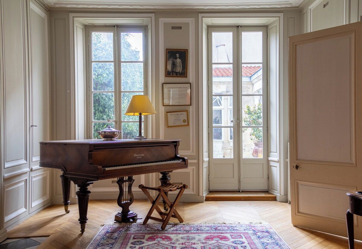 Un piano de cola en una habitación con ventanas amplias, una lámpara y diplomas enmarcados en la pared.