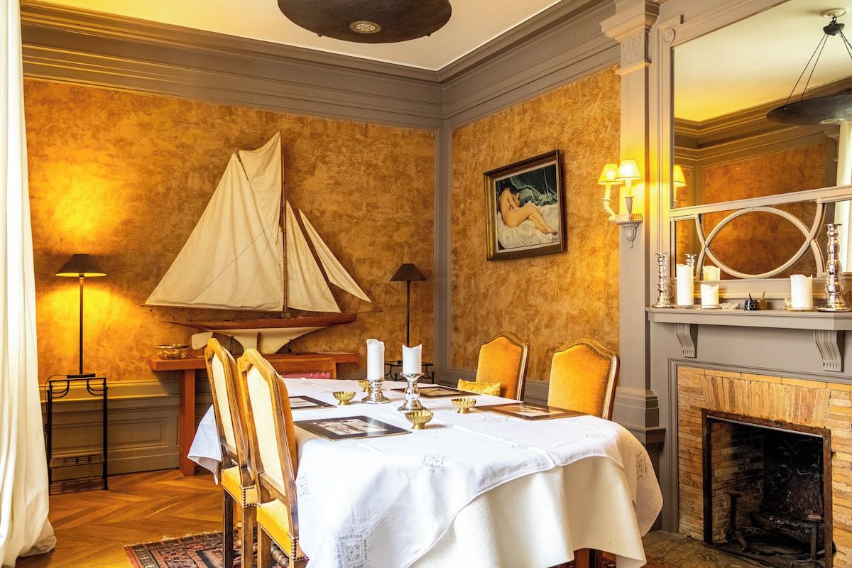 A dining room with a table set for four, a fireplace, and a painting on the wall.