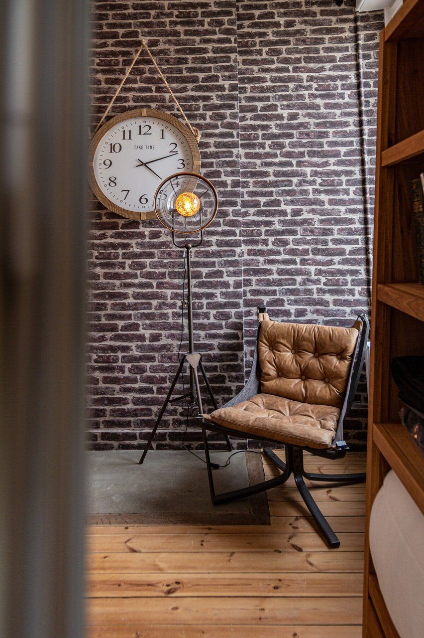Una habitación con pared de ladrillo, un reloj de pared redondo, una lámpara de pie, un sillón de cuero y una estantería.