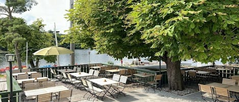Un espace extérieur aménagé avec des tables et des chaises, un grand arbre et une vue sur l’eau.