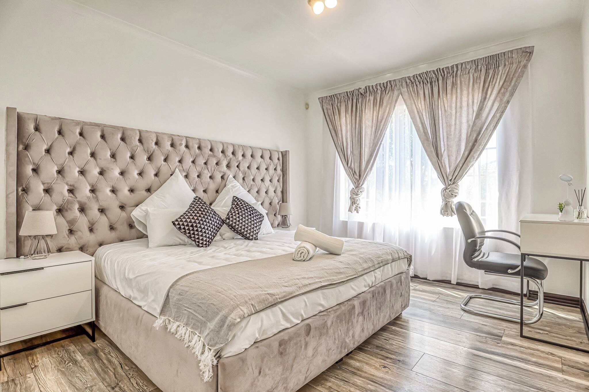 A bedroom with a tufted headboard, a bed with white bedding and patterned pillows, a bedside table with a lamp, a desk with a chair, and a window with curtains.