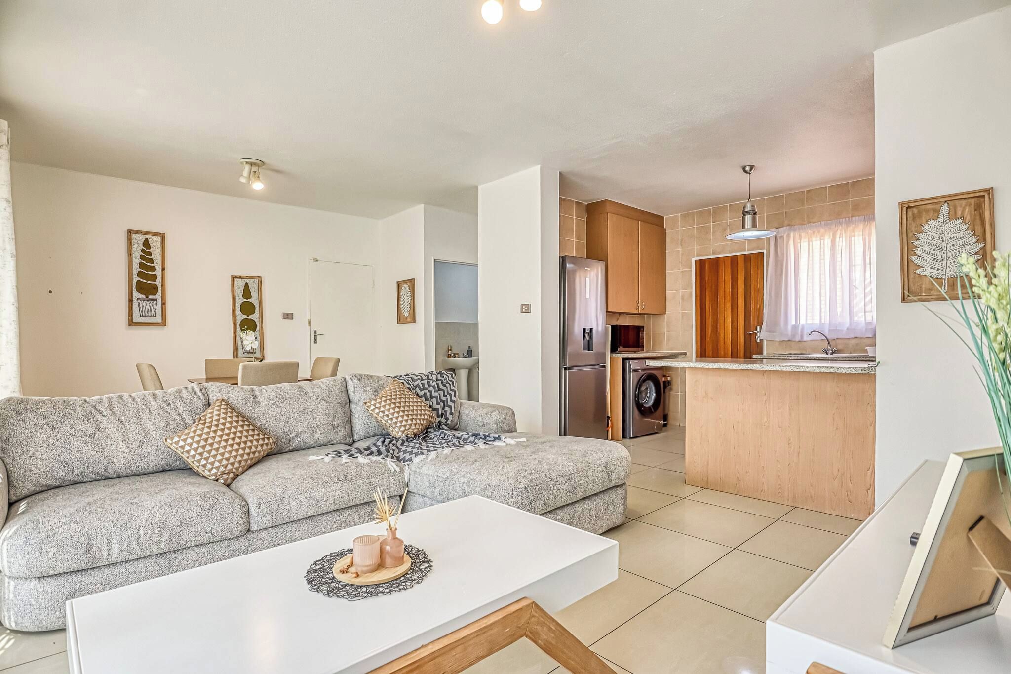 A modern living room with a grey sectional sofa, a white coffee table, and a kitchen area with wooden cabinets and a stainless steel refrigerator.