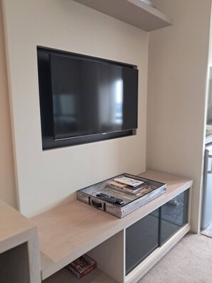 A flat-screen TV mounted on a wall above a wooden console with a storage box and books. - Sandton Studio with Views, Pool, Gym & Parking (Sandton)