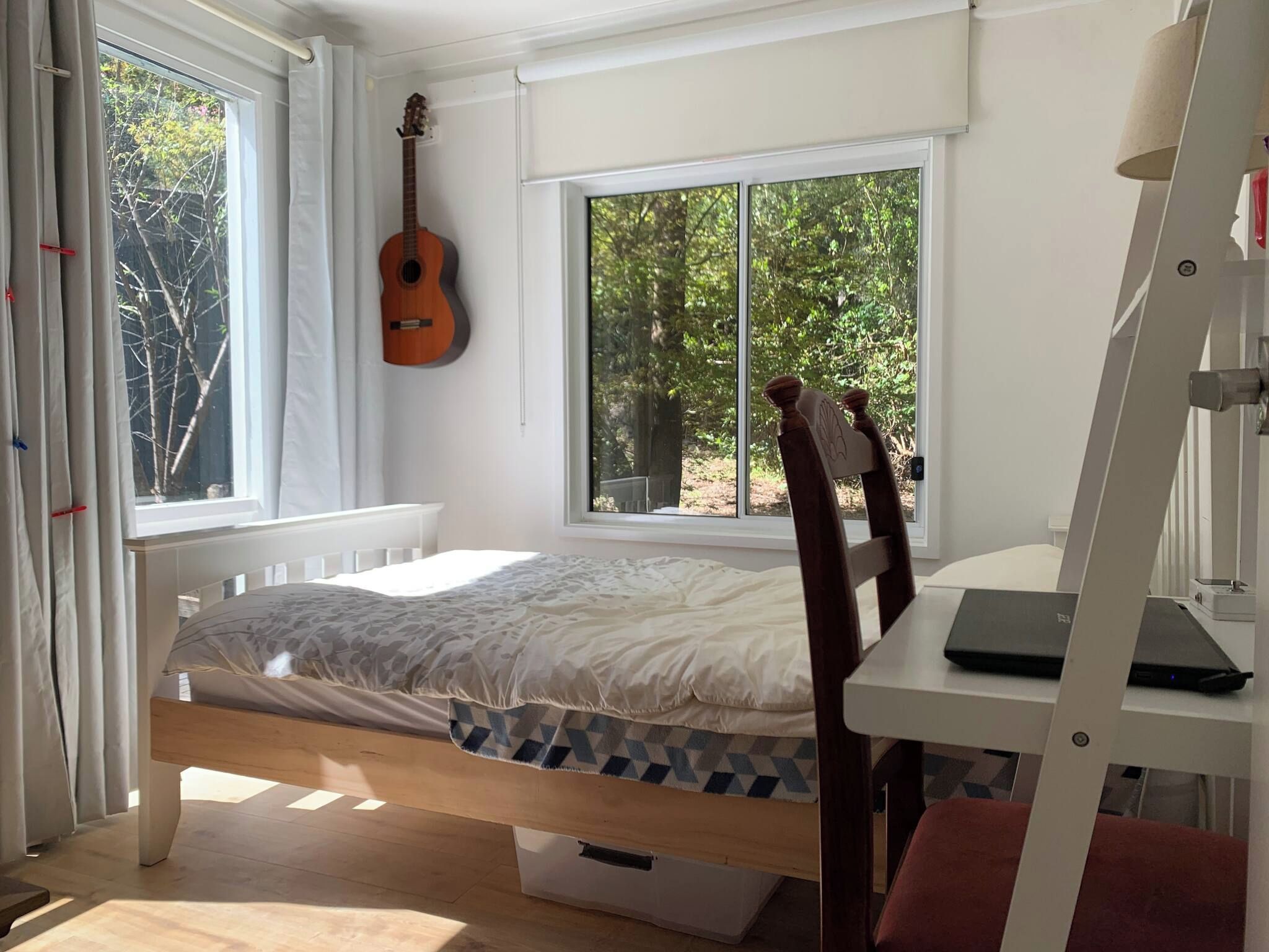 A compact room with a bed, a desk, a guitar, and a window overlooking greenery.