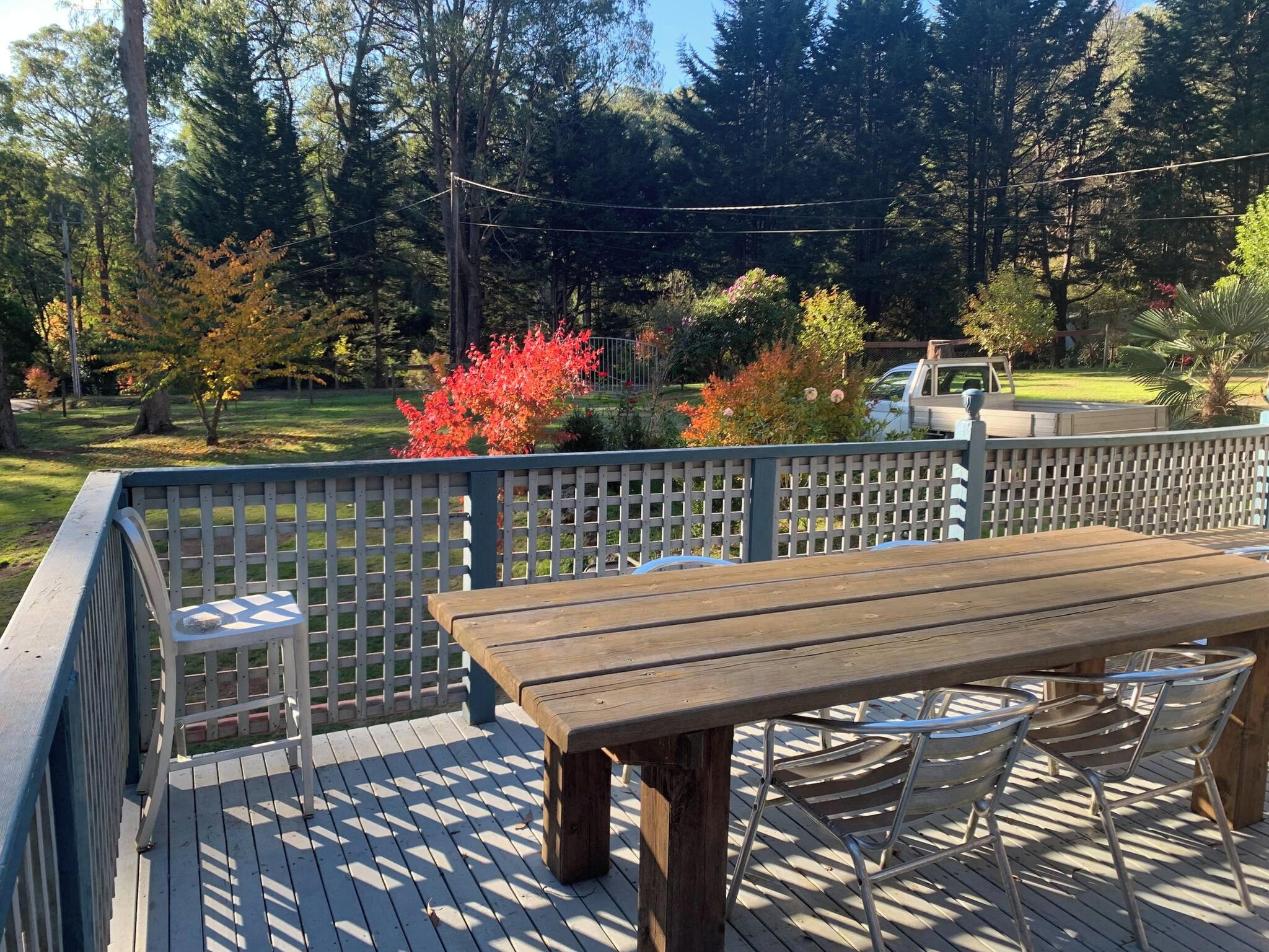 A wooden deck with a table and chairs, surrounded by a white fence, overlooking a garden with trees and a parked vehicle.