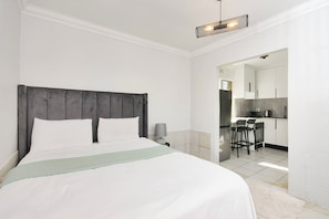 Een slaapkamer met een bed, een nachtkastje, een lamp en uitzicht op een keuken.