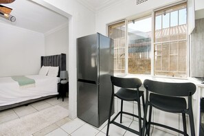 Ein kleines Zimmer mit einem Bett, einem Kühlschrank, zwei Stühlen und einem Fenster.