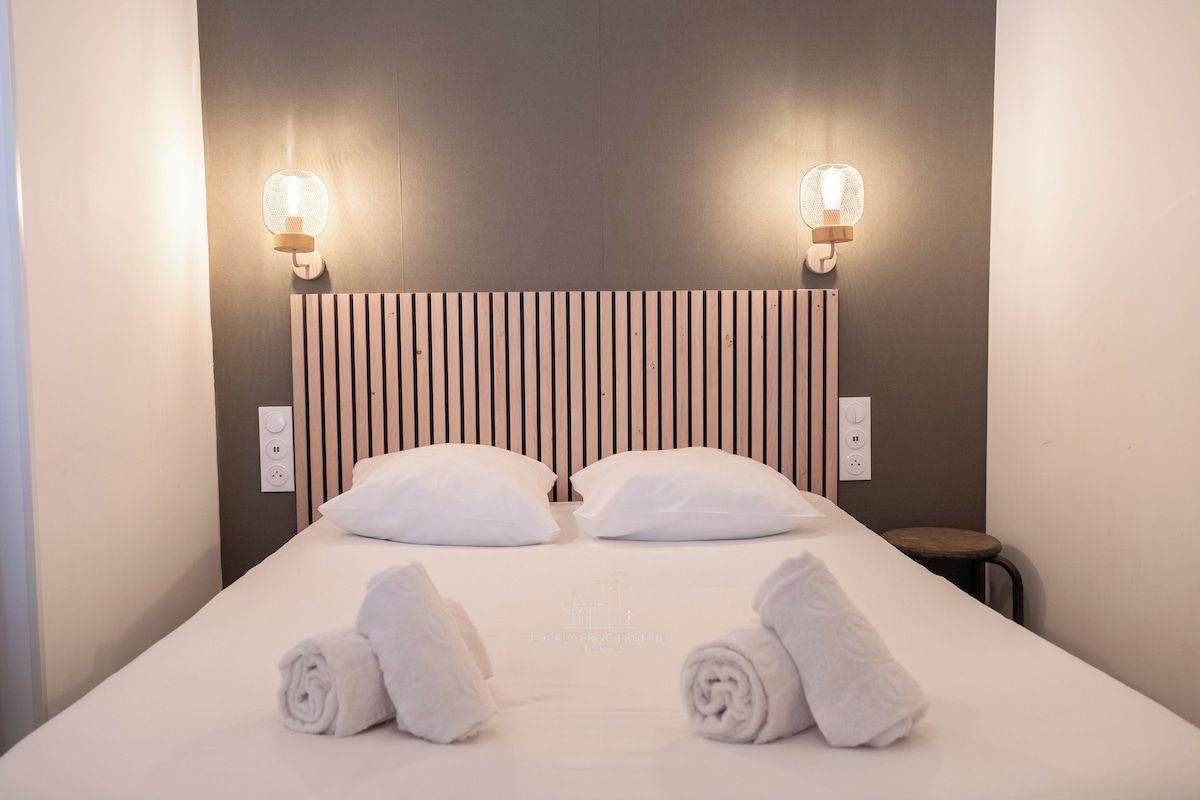 Habitación de hotel con cama, dos lámparas de pared, cabecera de madera y dos toallas enrolladas sobre la cama.