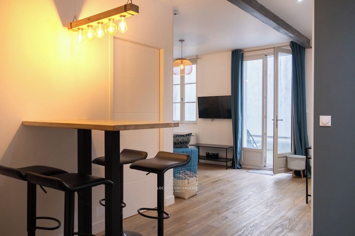 A modern living room with a wooden floor, a dining area with bar stools, a TV mounted on the wall, and a large window with curtains.