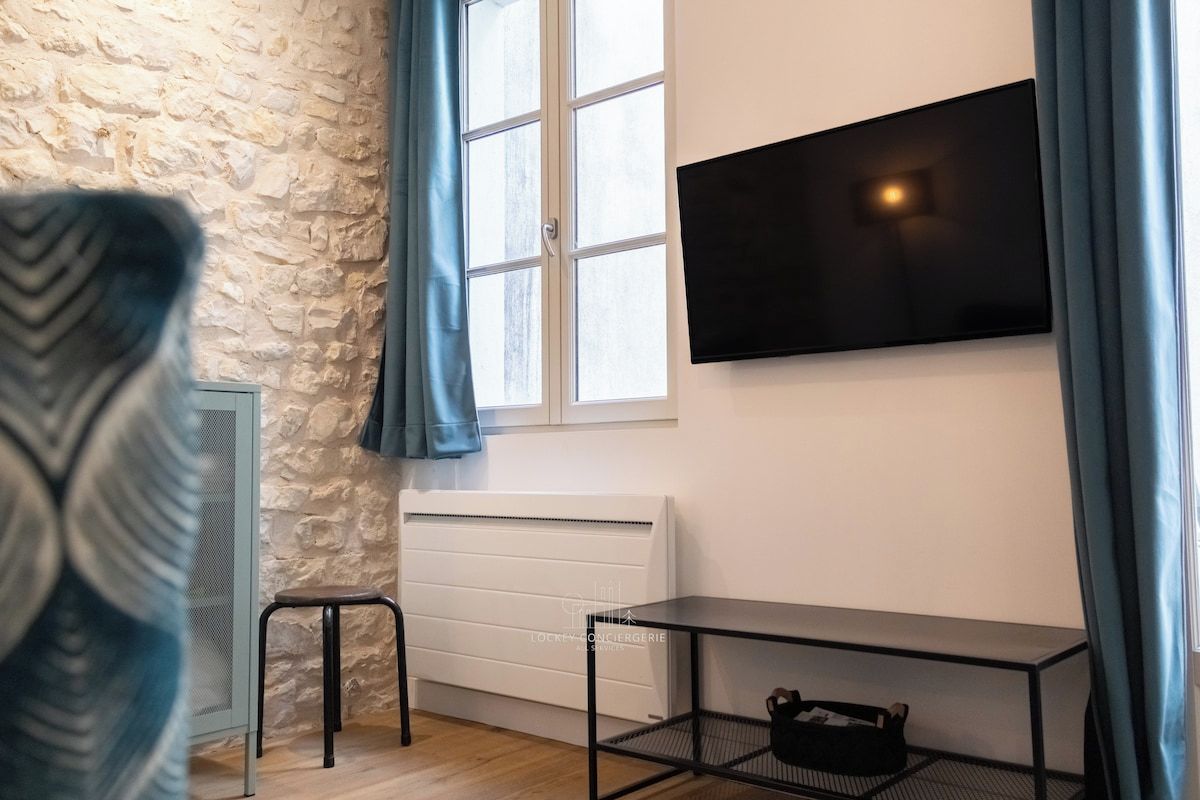 A room with a stone wall, a window with curtains, a radiator, a TV mounted on the wall, a small stool, and a metal table with a bag on it.