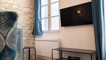 A room with a stone wall, a window with curtains, a radiator, a TV mounted on the wall, a small stool, and a metal table with a bag on it.