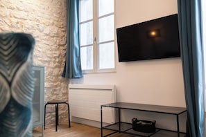A room with a stone wall, a window with curtains, a radiator, a TV mounted on the wall, a small stool, and a metal table with a bag on it. - Large Apartment with Patio - Steps from the Harbor (La Rochelle)