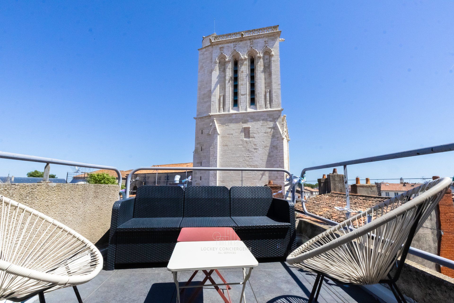 Una terrazza sul tetto con mobili in vimini, un tavolo e vista su una torre storica.