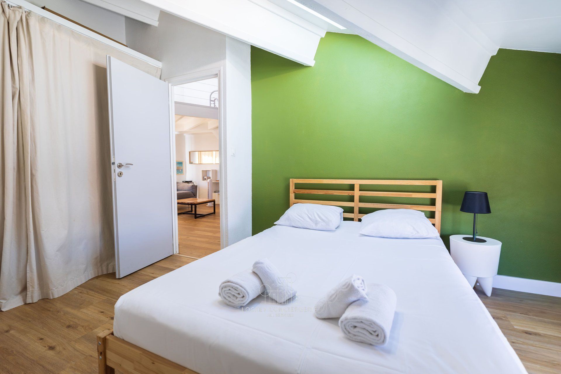 Una camera d'albergo con un letto, un comodino, una lampada e vista su un corridoio.