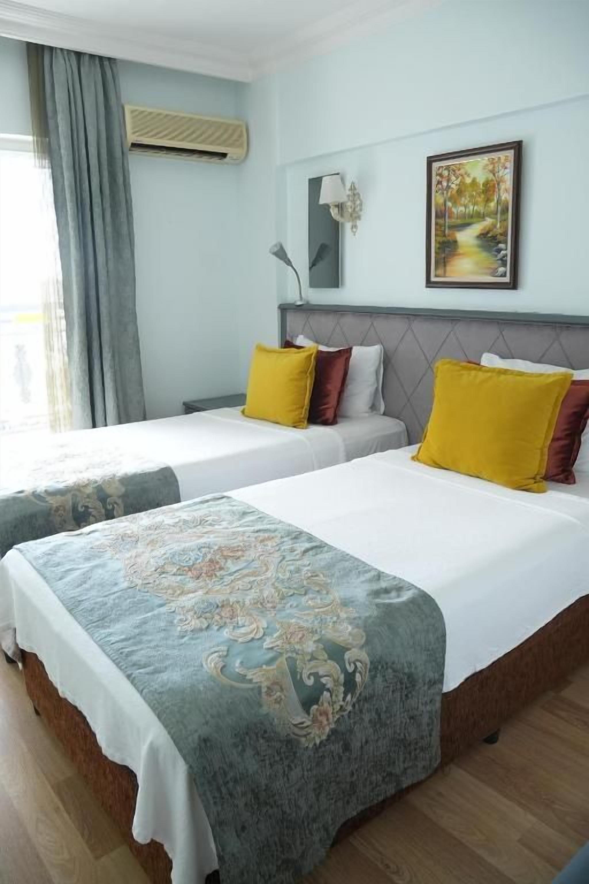 Habitación de hotel con dos camas, un ventanal con cortinas, un cuadro en la pared y una mesita de noche con lámpara.