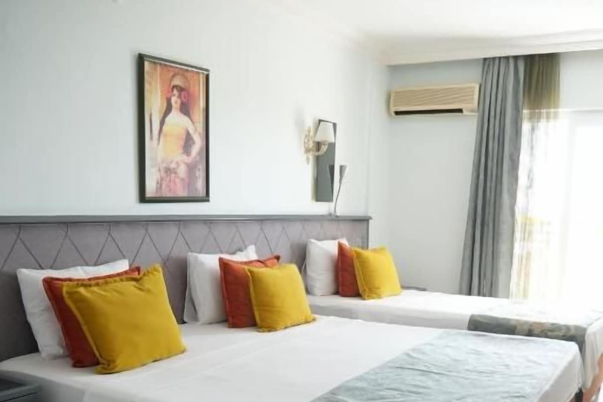 Quarto de hotel com duas camas, mesa de cabeceira, luminária na parede e janela com cortinas.