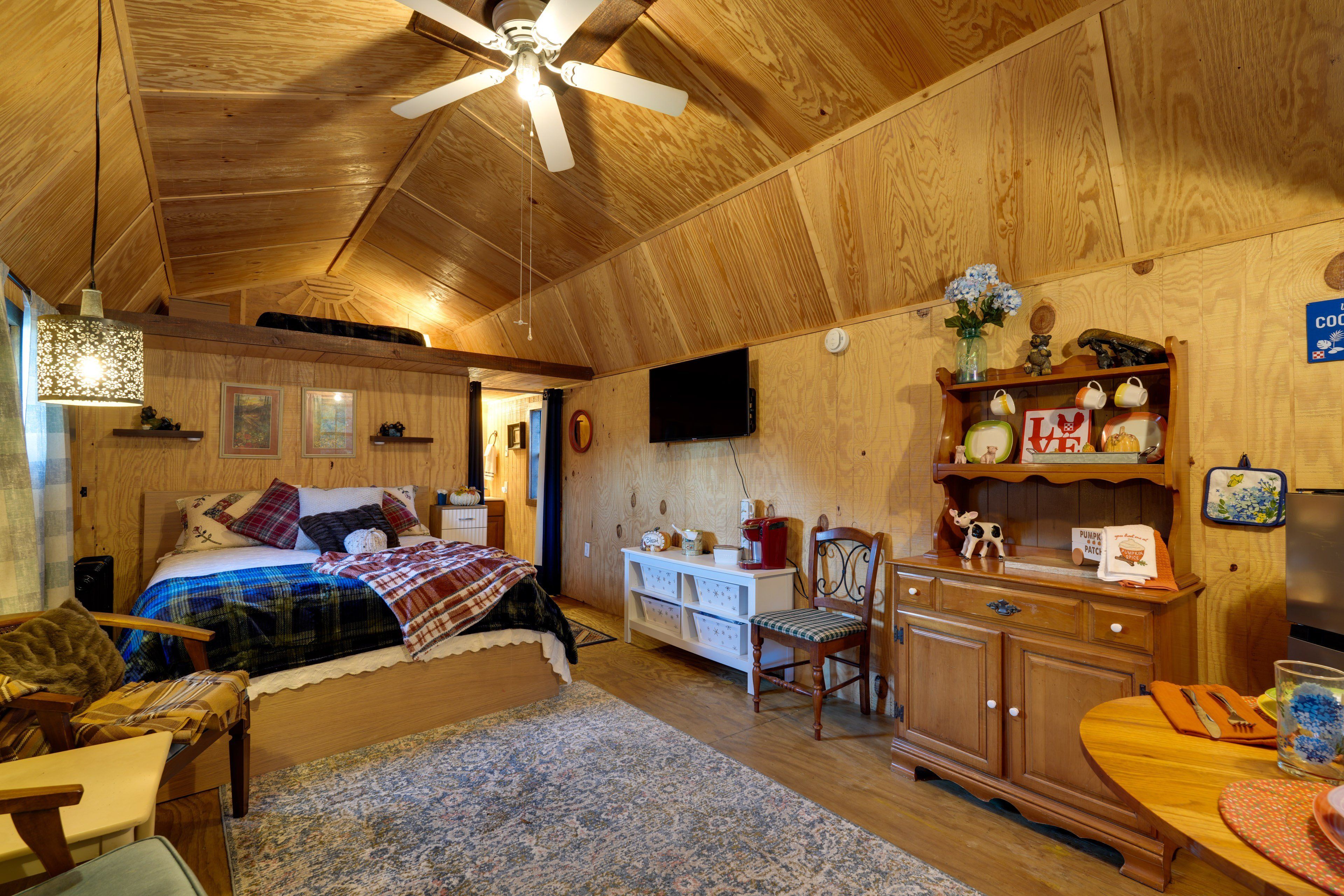 Een knus ingerichte kamer met een bed, bureau, stoel, televisie en plafondventilator.