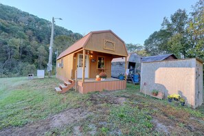 A wooden cabin with a porch, a small shed, and a utility box in a grassy area with trees in the background.