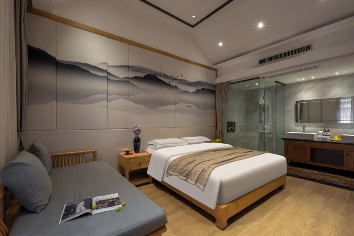 A modern bedroom with a large bed, bedside tables, a reading area, and a bathroom with a glass shower.
