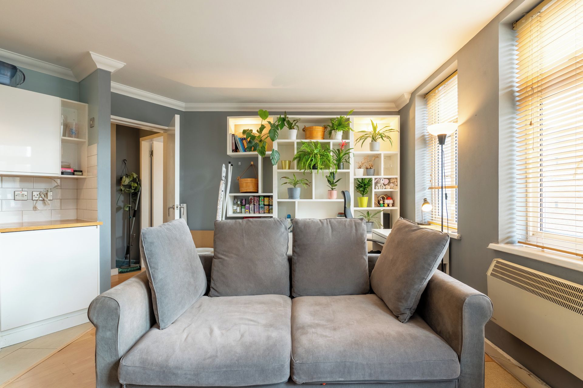 A living room with a grey sofa, a white kitchen in the background, and a window with blinds.