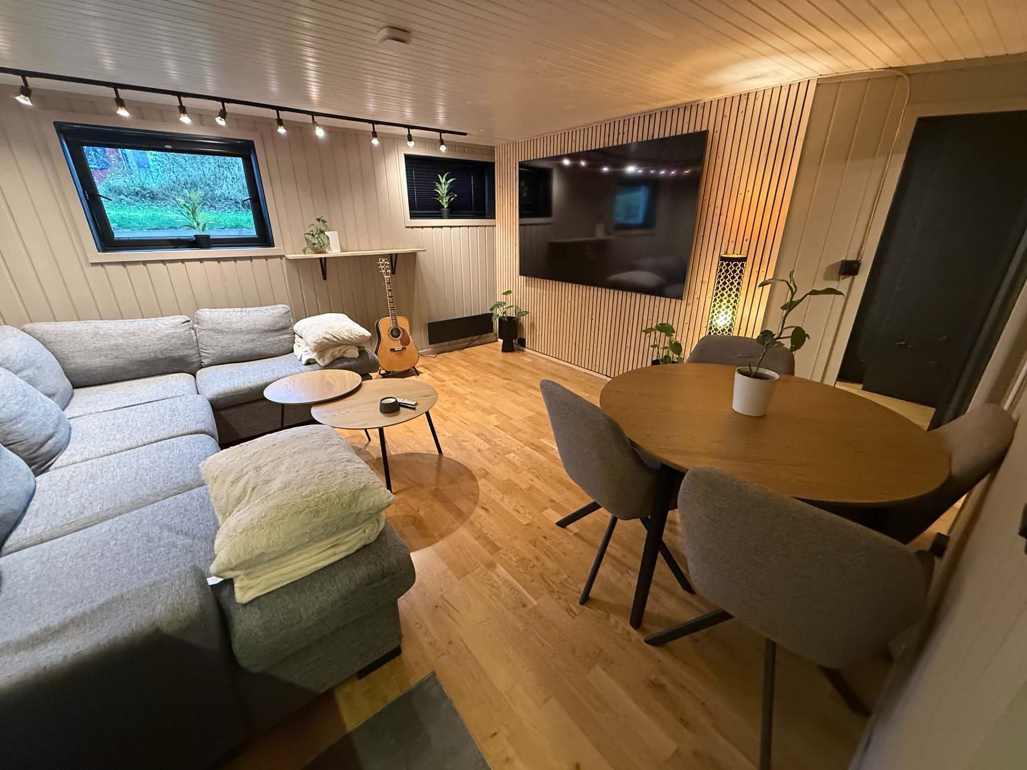 Un salon moderne comprenant un canapé, une table basse, une table à manger avec des chaises, une guitare et un téléviseur fixé au mur.