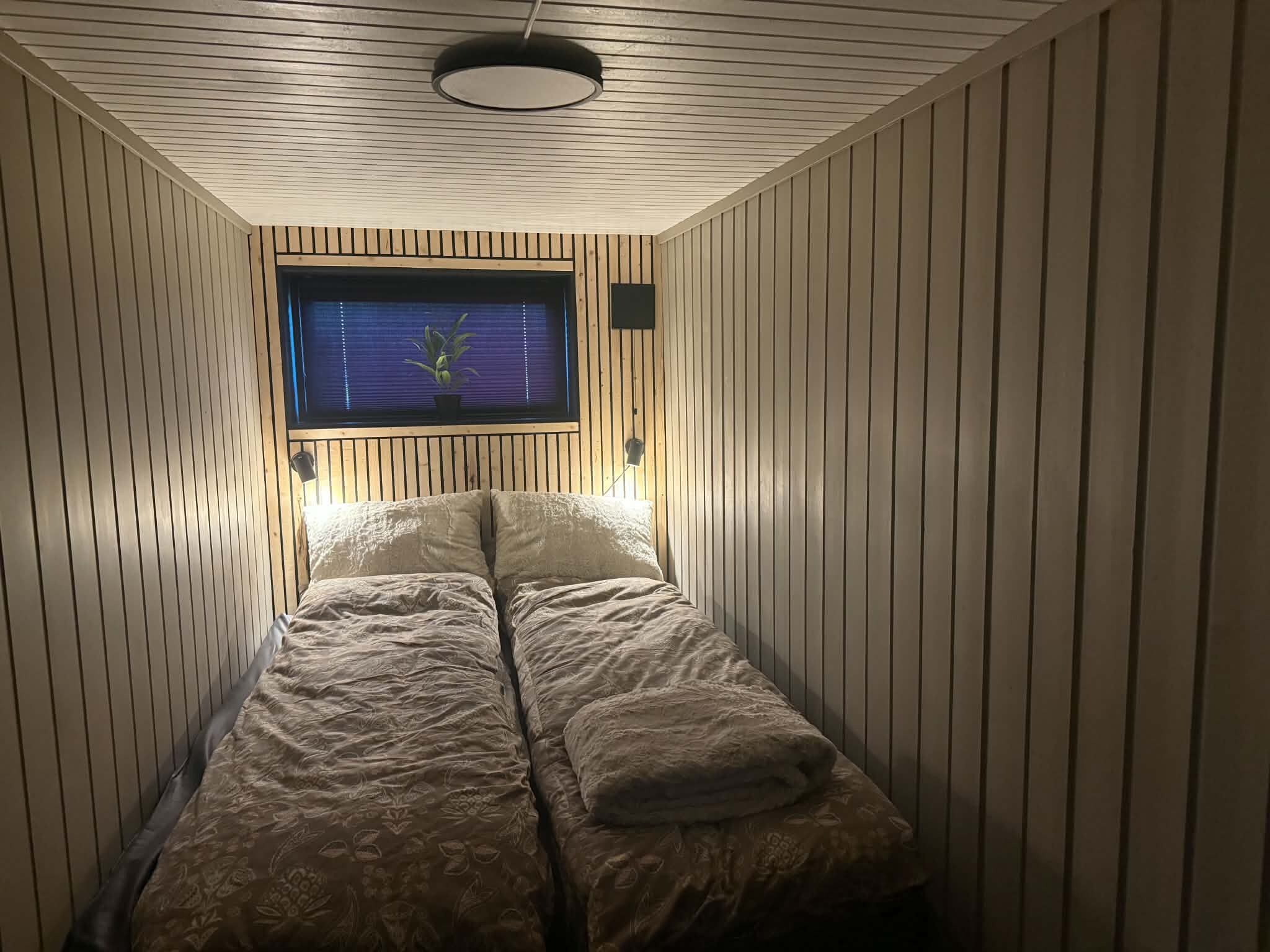 A narrow room with a single bed, wooden paneling on the walls, a window with blinds, and a ceiling light fixture.