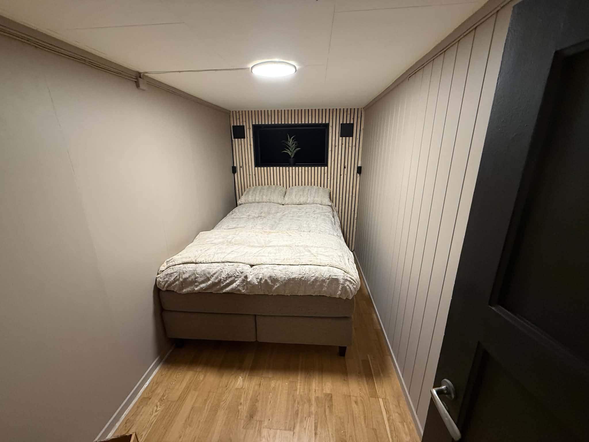 A small, narrow room with a single bed, a window with a plant, and a door.