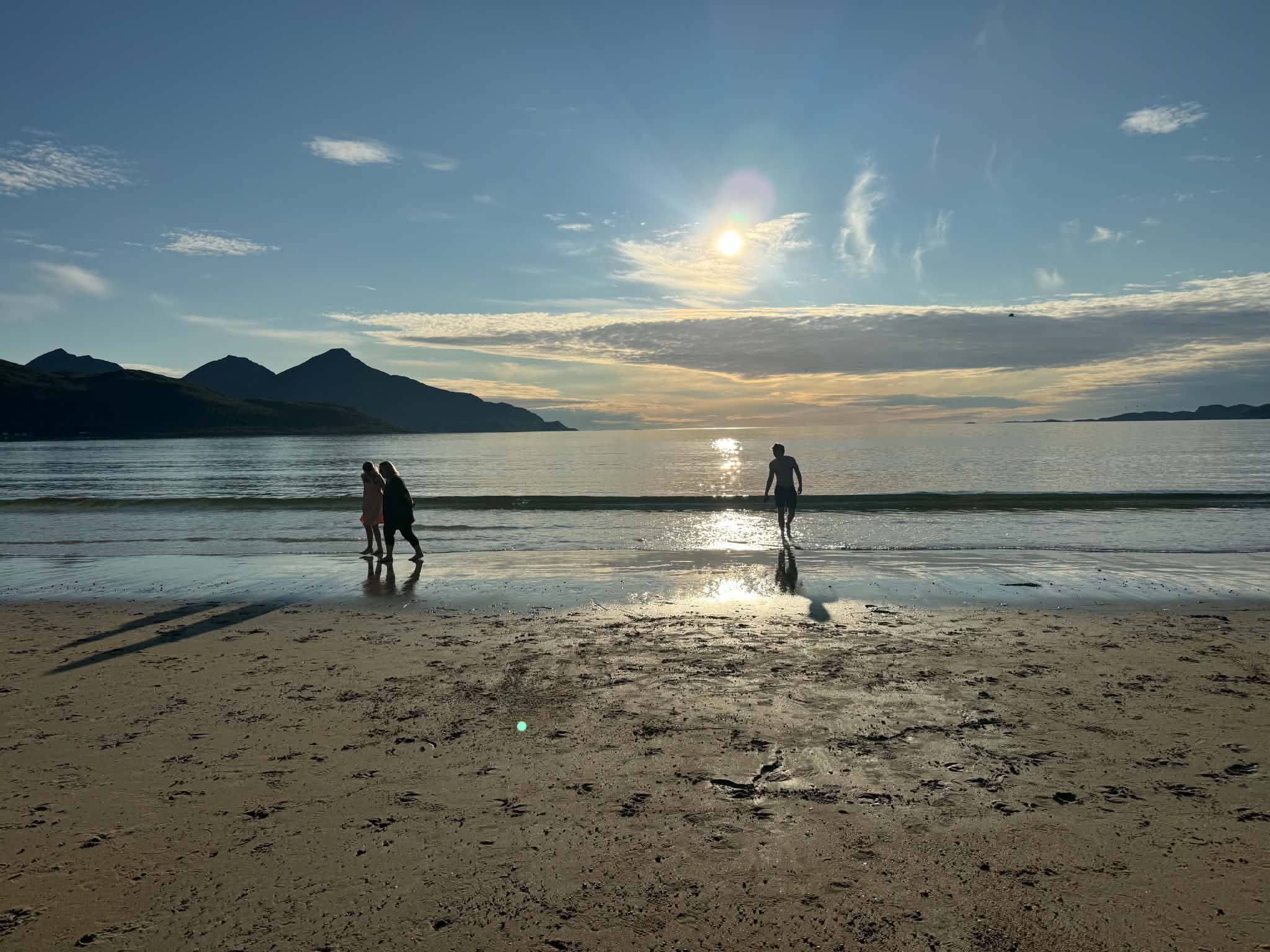 A beach with a calm sea, mountains in the distance, and a clear sky.